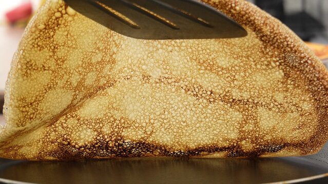Spatula lifting a pancake, revealing underside texture. The action depicts the turning process during cooking, showcasing the crispy golden exterior of the blin contrasted with the softer interior.
