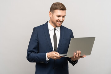 Successful man smiling broadly, looking at laptop screen. Satisfied facial expression, good offer, transaction, business video call. Studio portrait businessman with computer, isolated on gray wall.