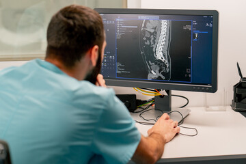 A radiologist sits at a table behind a computer monitor and examines a magnetic resonance imaging...