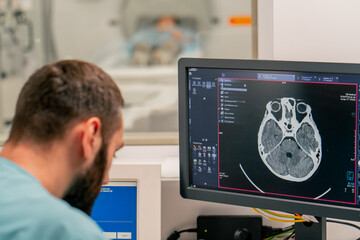 A radiologist sits at a table behind a computer monitor and examines a magnetic resonance imaging image during an examination of patient - obrazy, fototapety, plakaty