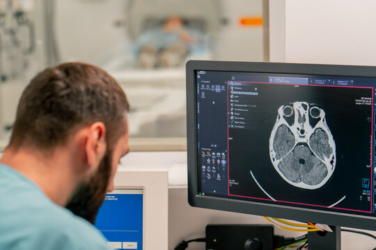 A radiologist sits at a table behind a computer monitor and examines a magnetic resonance imaging image during an examination of patient