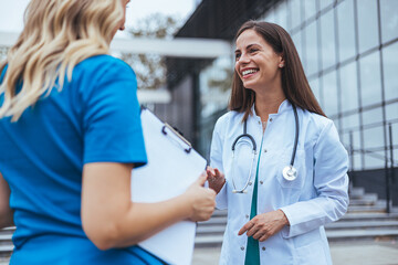 Two female doctor walk down an elevated walkway together. One wears a lab coat and the other wears scrubs. Female healthcare professionals walk and talk together