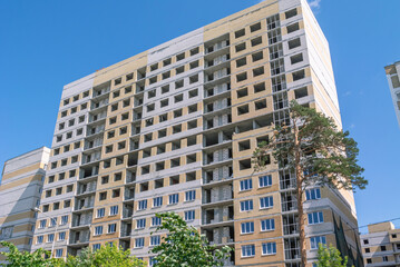 A pine tree grows in front of the house under construction. The high-rise residential building is lined with decorative bricks. The frame of a house under construction without window frames.