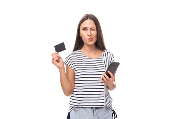 young positive brunette lady with straight hair in glasses dressed in a striped t-shirt uses a credit card and a smartphone on a white background with copy space