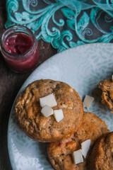 White chocolate chip cookies on a plate with raspberry jam jar on blue lace napkin on top of wooden rustic background