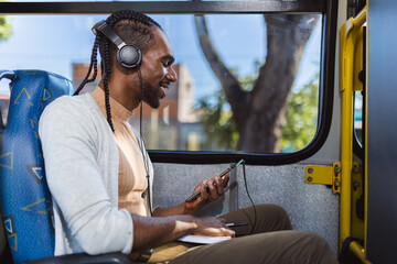 Jovem homem negro, usando fones de ouvido, ouvindo musica em seu smartphone, durante trajeto de onibus.