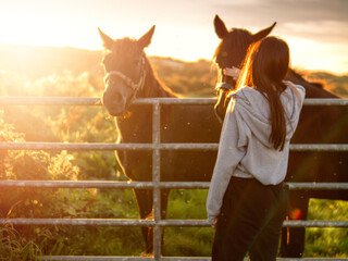 Obraz premium Teenager girl looking at dark horses by a metal gate to a field at stunning sunset. Warm sunshine glow. Selective focus. Light and airy soft look. Connection to nature concept. Rural area.