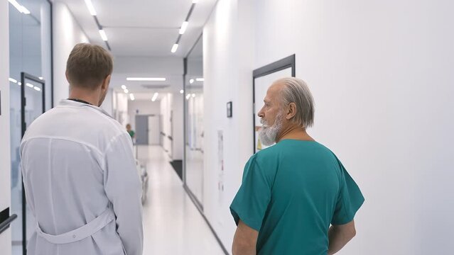 The chief physician, a surgeon in surgical attire, with a young doctor in a white coat, walks along the corridor of a modern clinic and discusses, gesticulating with his hands.