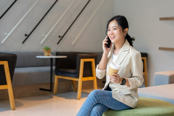 Cheerful Asian woman uses smartphone to work while drinking coffee in living room at home.