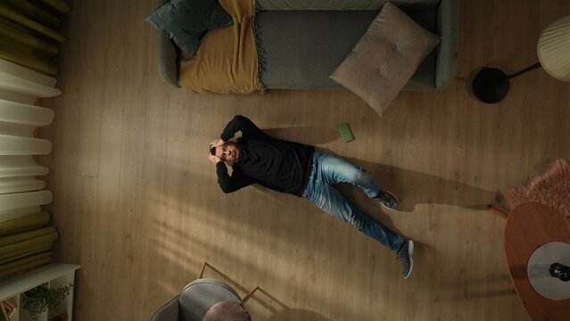 Top view of a shocked man lying on the living room floor next to a smartphone. The man is lying there clutching his head.