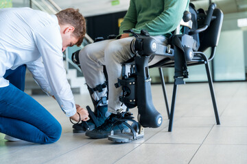 Mechanical exoskeleton. Physiotherapy in a modern hospital: Physiotherapist placing the tapes on the knees of the disabled person to place his robotic skeleton - obrazy, fototapety, plakaty