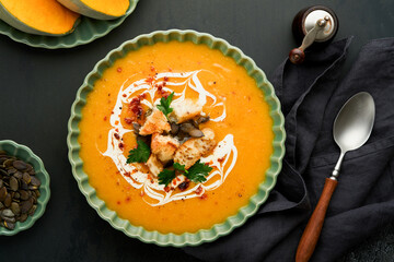 Pumpkin and carrot cream soup with herbs, seasonings and seeds in bowl on black wooden background in rustic style. Thanksgiving traditional autumn pumpkin cream soup. Top view.