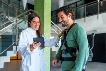 Mechanical exoskeleton, female physiotherapist doctor talking and smiling with tablet with disabled person with robotic skeleton, physiotherapy in modern hospital, futuristic physiotherapy
