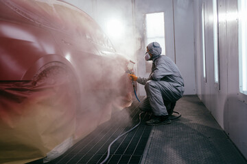 Automobile repairman painter in protective glove with airbrush pulverizer painting car body in paint chamber. Automotive paint services, quality auto body shop concept. - obrazy, fototapety, plakaty