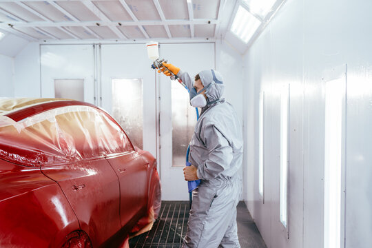 Automotive paint services, quality auto body shop concept. Real shot of man of professional car painter with protective clothing standing next to car in auto paint room and with gun