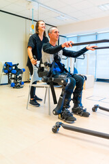 Mechanical exoskeleton. Female physiotherapy doctor assistant lifting disabled person with robotic...