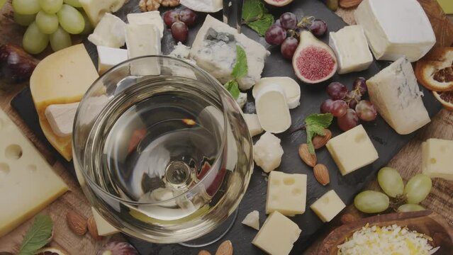Glass of white wine with variety of sliced cheeses with fruits, mint, nuts and cheese cutting knives slowly rotate in the frame. Wonderful cheese and wine background for your projects.