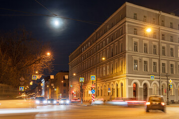 Evening cityscape. Cars drive through the streets of the city at night. Bright moon in the sky. Long exposure. Blurred traces from car headlights. Historical center of St. Petersburg, Russia.