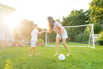 Young mother and son giving five during soccer game in the garden. Happy family playing football, having fun together. Fun Playing Games in Backyard Lawn on Sunny Summer Day. Motherhood, childhood - obrazy, fototapety, plakaty