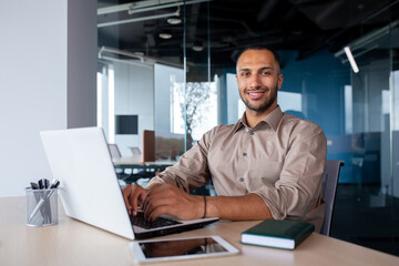 Fototapeta na wymiar Attractive african american man sitting at desk in front of laptop in office, programmer working in modern environment, tapping on keyboard and smiling at camera.
