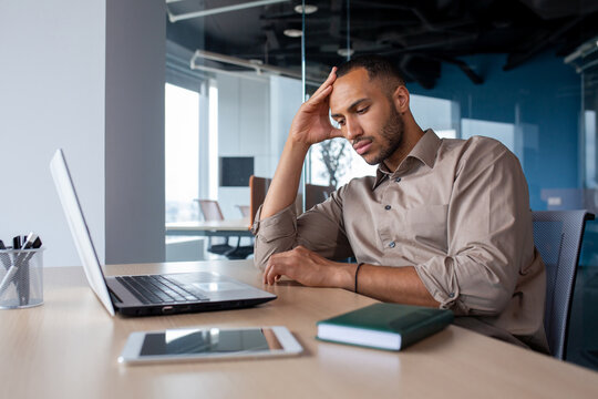 Upset tired young african american man sitting at desk with laptop in office, tired of work holding hand on head, suffering from headache, bored in office.