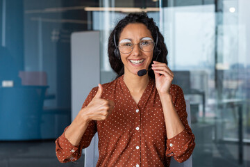 Happy indian woman in headset showing thumb up in office. Satisfied curly young businesswoman in glasses is happy to work in modern room, smiling at camera.