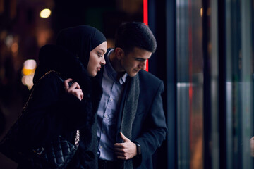 Fototapeta premium Happy multicultural business couple walking together outdoors in an urban city street at night near a jewelry shopping store window.