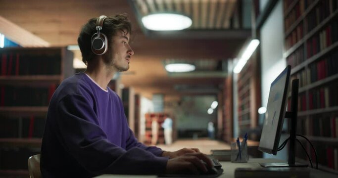 Young Male Student in Purple Jumper Working on His Master's Thesis on a Desktop Computer. Smart Man Learning a Lecture Online, Looking Through a Presentation in a Library with Book Shelves