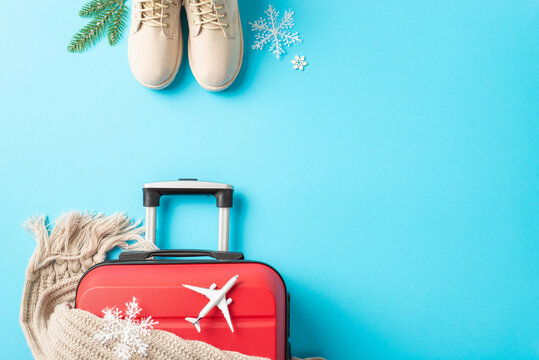 New Year's Flight Preparations: top view image of a red suitcase, miniature plane, winter essentials – boots, scarf, garnished with fir twigs and snowflakes against a vibrant blue backdrop