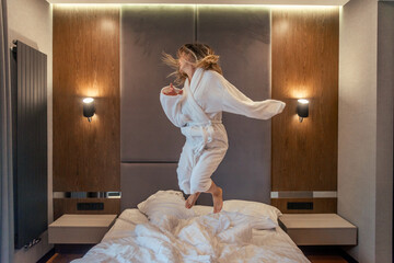 Woman having fun and jumping with bathrobe on bed in hotel room - obrazy, fototapety, plakaty
