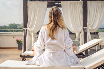 Woman in white bathrobe lying on sofa and relaxing with orange cocktail at the roof.