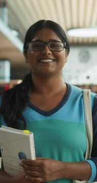 Vertical Screen: Portrait of a Cheerful Indian Student Standing in a Traditional Public Library. Young South Asian Female Looking at Camera and Smiling. Scholar Holding Academic STEM and Math Textbook