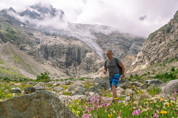 Fototapeta premium Panorama of a glacier in the mountains. Granite stones in the mountains. A man is walking, and a glacier is visible behind. View of the mountains, clouds and flowers. Hiking in the mountains. 