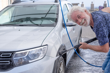 Smiling white-haired senior man washing his car in a self-service car wash station using high...