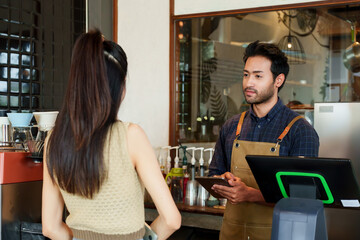 Businessman, cafe owner, multiracial male holding tablet, taking coffee and bakery orders from female Asian customer cream-colored shirt. front of cash register counter of small cafe small business.