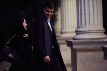 Happy multicultural business couple walking together outdoors in an urban city street at night near a jewelry shopping store window.