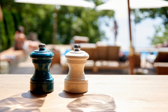 Salt and pepper shakers next to each other. They are isolated and stand by themselves, on a table in a restaurant, outside. A chic restaurant with sun loungers and a swimming pool is depicted in the