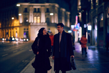 Happy multicultural business couple walking together outdoors in an urban city street at night near a jewelry shopping store window.