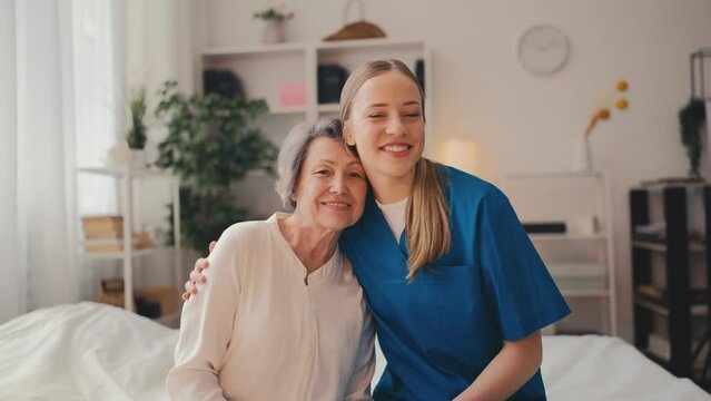 Portrait of friendly nurse hugging smiling senior woman patient at hospital room