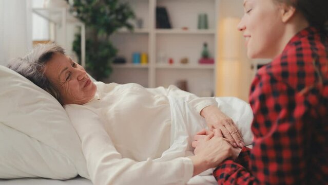 Young woman talking and holding hand of smiling grandmother resting in bed