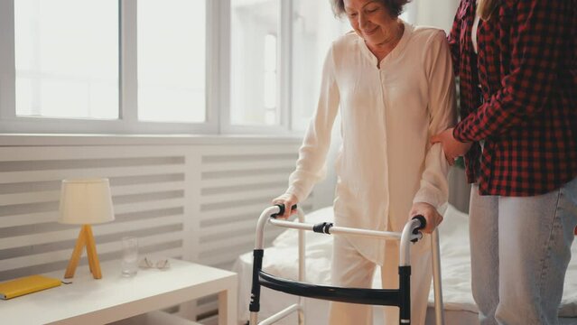 Caring young woman helping elderly grandmother to use walking frame at home