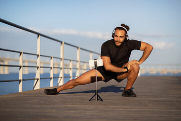 fitness guy in deep lunge, streaming exercise via smartphone outdoors