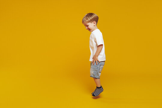 Horizontal image of side view full body stylish little kid boy in white t-shirt and striped socks, jumping while smiling against yellow background.