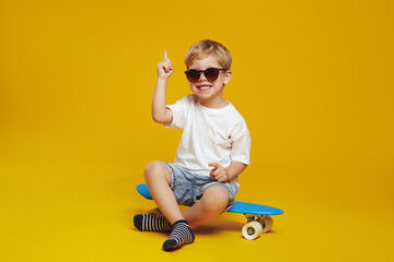 Adorable little boy in white tshirt and sunglasses, sitting on modern skateboard while looking at camera and pointing up, against yellow background. - obrazy, fototapety, plakaty