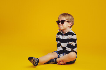 Full body of little kid boy wearing trendy sunglasses and striped shirt looking away while sitting...