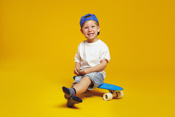 Full body length excited kid boy wearing trendy blue cap and white t-shirt, smiling while sitting on modern skateboard against yellow background. © Davidovici