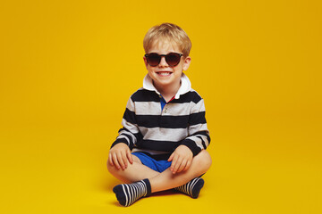 Cute kid boy wearing trendy sunglasses and striped shirt smiling while sitting with crossed legs on...