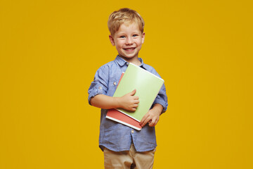 Excited little genius child with blonde hair in blue shirt holding textbooks and smiling happy while standing against blue background, elementary school concept.