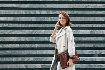 Side view of stylish middle-aged businesswoman in coat. Middle-aged female talking on mobile phone while walking.