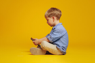 Side view of focused little boy sitting on studio floor with crossed legs while playing addictive games on smartphone, isolated over yellow background. Gadget addiction. - obrazy, fototapety, plakaty